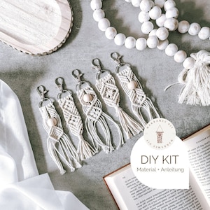 Könnte beinhalten: Fünf Makramee-Schlüsselanhänger mit Holzperlen und Quasten. Die Schlüsselanhänger sind beige und weiß und liegen auf einer grauen Oberfläche. Das Bild zeigt außerdem ein Buch, ein Tablett aus Holz und eine weiße Perlenkette.