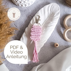 May include: A pink macrame keychain with a silver clasp. The keychain features a woven rectangular design and a tassel. The image also includes the text "PDF & Video Anleitung".