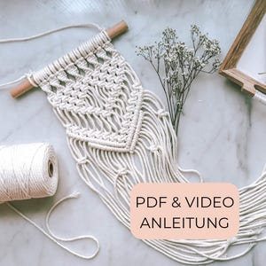 以下が含まれることがあります： 木製のダボが付いたクリーム色のマクラメの壁掛け。手作りのテキスタイルは、複雑な結び目のパターンとフリンジが特徴です。白い綿コードのロールが見え、テキスト「PDF＆VIDEO ANLEITUNG」が表示されています。
