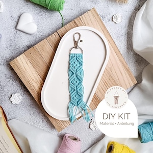 Könnte beinhalten: Ein türkisfarbener Makramee-Schlüsselanhänger mit silbernem Verschluss, präsentiert auf einem weißen Tablett auf einem Holzbrett. Der Schlüsselanhänger hat ein geometrisches Muster und Fransen. Ein DIY-Kit-Etikett ist sichtbar.