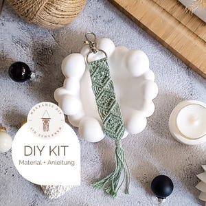 Könnte beinhalten: Ein salbeigrüner Makramee-Schlüsselanhänger mit silbernem Verschluss und Quaste. Der Schlüsselanhänger wird auf einer weißen, dekorativen Schale präsentiert. Das Bild enthält ein DIY-Kit-Etikett mit dem Text "DIY KIT Material + Anleitung".
