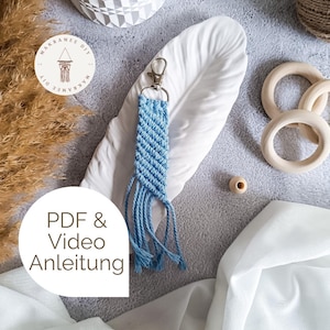 Puede incluir: Un llavero de macramé azul hecho a mano con un cierre plateado. El llavero presenta un diseño tejido con flecos colgantes. La imagen también incluye anillos de madera, una cuenta de madera y el texto "PDF & Video Anleitung".