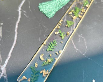 May include: A rectangular resin bookmark with a gold border, featuring green fern leaves and gold flakes encased in clear resin. A green tassel is attached to the top. The Indigo Resin Art logo is visible in the bottom right corner.