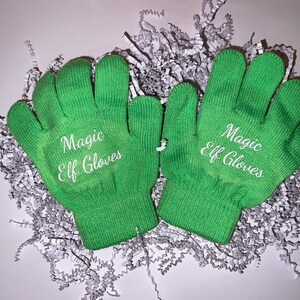 May include: A pair of bright green knit gloves with the words "Magic Elf Gloves" embroidered in white. The gloves are displayed on a bed of white shredded paper.
