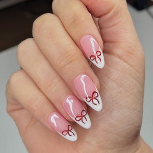 Puede incluir: Una mano con uñas largas en forma de almendra. Las uñas presentan un diseño de manicura francesa con base rosa, puntas blancas y pequeños acentos de lazo rojo. Las uñas están bien cuidadas y pulidas, mostrando una estética delicada y femenina.