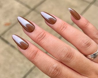May include: Close-up of a hand with long, almond-shaped nails painted a rich brown color with a glossy finish.