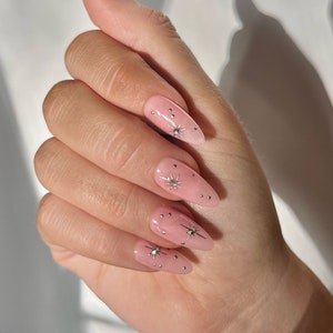 May include: A hand with long, almond-shaped nails painted a light pink colour. Each nail is decorated with small silver stars and a larger silver star in the centre.
