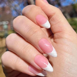 Peut inclure: Gros plan sur une main avec de longs ongles en amande. Les ongles sont peints d'un vernis rose clair et ont une pointe blanche avec une finition texturée et scintillante.