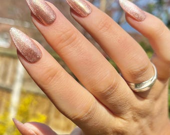 May include: Close-up of a hand with long, almond-shaped nails painted with a shimmery rose gold nail polish.