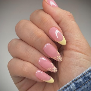 Puede incluir: Una mano con uñas artificiales largas en forma de almendra. Las uñas están pintadas con una base rosa claro, una punta amarilla y un diseño de media luna blanca. Algunas uñas presentan pequeños acentos de margaritas blancas. Las uñas son brillantes y están bien cuidadas.