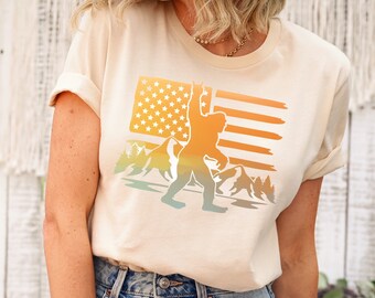 May include: A cream colored t-shirt with a graphic of a Bigfoot standing in front of a sunset-colored American flag. The graphic also includes mountains and trees.