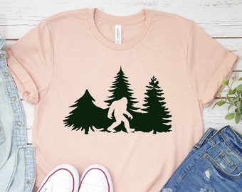 May include: A pink t-shirt with a black silhouette of Bigfoot walking through a forest of pine trees.