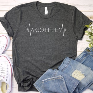 Pode incluir: Uma camiseta cinza escura com um gr&aacute;fico branco de um batimento card&iacute;aco com a palavra "COFFEE" dentro. A camiseta est&aacute; colocada sobre uma superf&iacute;cie de madeira branca com um par de t&ecirc;nis brancos e uma cal&ccedil;a jeans azul.