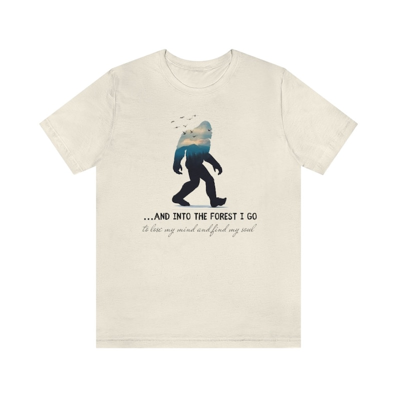 Peut inclure: Un t-shirt beige avec une silhouette de Bigfoot marchant dans une for&ecirc;t. Le texte dit "...AND INTO THE FOREST I GO to lose my mind and find my soul."