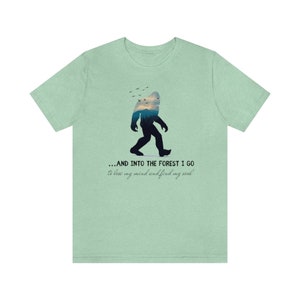 Peut inclure: Un t-shirt vert clair avec une silhouette de Bigfoot marchant dans une sc&egrave;ne de for&ecirc;t. Le texte dit "...AND INTO THE FOREST I GO to lose my mind and find my soul."