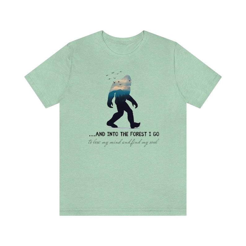 Peut inclure: Un t-shirt vert clair avec une silhouette de Bigfoot marchant dans une sc&egrave;ne de for&ecirc;t. Le texte dit "...AND INTO THE FOREST I GO to lose my mind and find my soul."