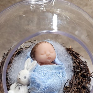 May include: A small, sleeping baby doll wrapped in a blue blanket is nestled in a bird's nest inside a clear glass ornament. A white stuffed bunny sits beside the doll.