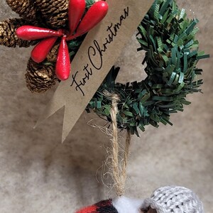 May include: A handmade Christmas bauble featuring a sleeping baby doll wrapped in a red and black plaid blanket and a white knitted hat. The bauble is hanging from a green wreath with a brown paper tag that says "First Christmas".