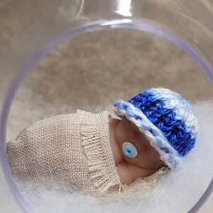 Könnte beinhalten: Eine kleine, handgefertigte Baby-Puppe, die einen blau-weißen Häkelhut und eine beige Wickeldecke trägt. Die Puppe liegt auf einem Bett aus weißer Watte in einer durchsichtigen Glaskuppel.