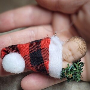 Könnte beinhalten: Eine kleine, schlafende Babypuppe, die einen rot-schwarzen karierten Hut trägt und einen grünen, belaubten Zweig hält. Die Puppe liegt auf dem Rücken mit geschlossenen Augen.