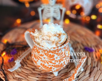 May include: A festive Halloween-themed image featuring a white and orange Talavera mug filled with whipped cream and a matching spoon. The mug and spoon have a floral design. The scene includes a skeleton, pumpkins, and string lights.