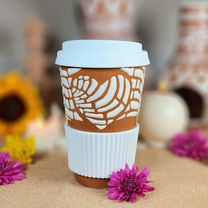 May include: A terracotta-colored coffee cup with a white, stylized bone design. The cup has a white lid and a white ribbed sleeve. The cup is decorated with a white bone design. The cup is surrounded by flowers.
