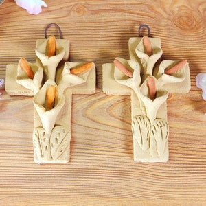 May include: Two beige ceramic crosses with calla lily designs. Each cross features a central vertical beam and a horizontal beam, with sculpted calla lilies and orange-colored stamens. The crosses have a loop for hanging.