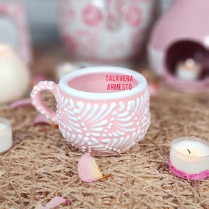 May include: A pink ceramic teacup with white floral detailing and the text "Talavera Armesto" inside. The teacup has a handle and is surrounded by candles and rose petals. The teacup is on a woven surface.