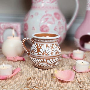 May include: A brown ceramic mug with white floral designs, a handle, and a white rim. The mug is surrounded by lit candles, pink rose petals, and decorative pink ceramic pieces, creating a cozy, romantic ambiance.