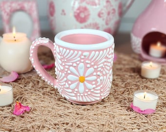 May include: A pink ceramic mug with a white daisy design. The mug has a white rim and a white handle. The mug is sitting on a brown woven surface.