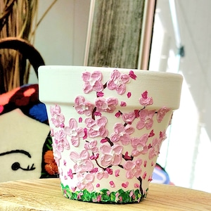 May include: A white ceramic flower pot decorated with pink cherry blossoms. The blossoms are textured and painted on the pot's exterior. Green grass and pink hearts are painted at the base of the pot. The pot sits on a wooden surface.