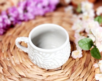 May include: White ceramic mug with a raised floral design. The mug has a rounded shape and a small handle.