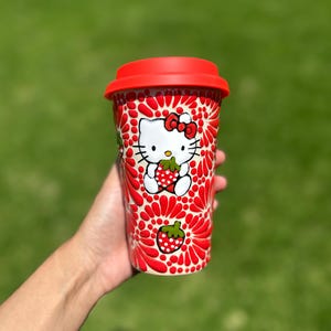 May include: A red travel mug featuring a Hello Kitty design holding a strawberry. The mug has a red lid and a pattern of red flowers and strawberries. The background is a blurred green.