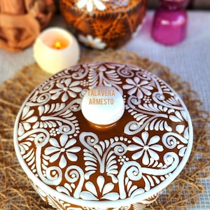 May include: A brown ceramic container with a white floral design and a white knob. The container has the text "TALAVERA ARMESTO" on the knob. The container is on a woven mat.