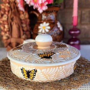 May include: A peach-colored ceramic dish with a lid, adorned with yellow and black butterfly designs and white floral patterns. The lid features a white knob. The dish sits on a woven mat, with the text "Casavera Armesto" visible on the side.