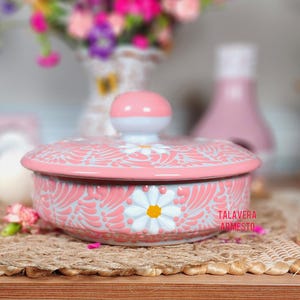 May include: A pink ceramic dish with a lid, featuring a white daisy design and a floral pattern. The dish is decorated with a light blue background and has the words "TALAVERA ARMESTO" on the side.