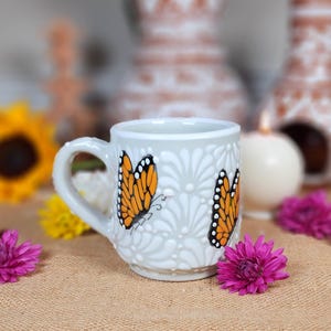 May include: White ceramic mug with a raised floral design and two hand-painted orange butterflies. The mug has a handle and is perfect for coffee or tea.