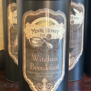 May include: Black cylindrical tea tins with decorative labels. The labels feature the text "Moon Honey Tea Co." and "Witches' Breakfast" with Assam Black Tea & Garden Herbs. The design includes a cauldron and a broom.