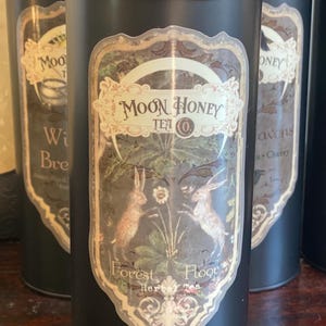 May include: A black metal tin with a label that says "Moon Honey Tea Co." and "Forest Floor Herbal Tea". The label features a whimsical illustration of two rabbits in a forest setting.