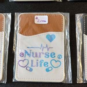 May include: Three white phone wallet cases with different designs. The first case has a butterfly and the word "nurse" in purple. The second case has a heart, a stethoscope, and the text "Nurse Life". The third case has a heart, a stethoscope, and the word "Nurse".
