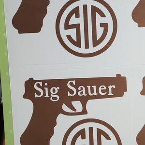 K&ouml;nnte beinhalten: Braune Vinyl-Aufkleber mit dem Text "Sig Sauer" in Pistolenform und einem kreisf&ouml;rmigen "SIG"-Logo. Die Aufkleber befinden sich auf wei&szlig;em Hintergrund, mit einer gr&uuml;nen Schneidematte.