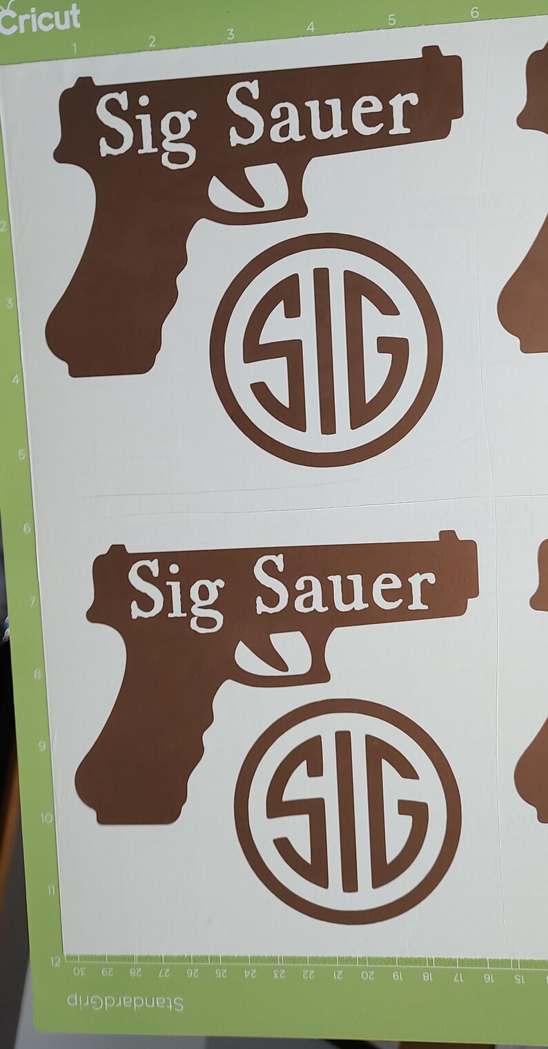 K&ouml;nnte beinhalten: Braune Vinyl-Aufkleber mit dem Text "Sig Sauer" in Pistolenform und einem kreisf&ouml;rmigen "SIG"-Logo. Die Aufkleber befinden sich auf wei&szlig;em Hintergrund, mit einer gr&uuml;nen Schneidematte.