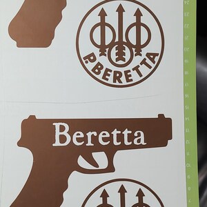 K&ouml;nnte beinhalten: Braune Vinylaufkleber mit dem Wort "Beretta" in Wei&szlig;, einer Pistouetten-Silhouette und einem kreisf&ouml;rmigen Logo. Das Logo enth&auml;lt das Wort "Beretta" und ein stilisiertes Design.