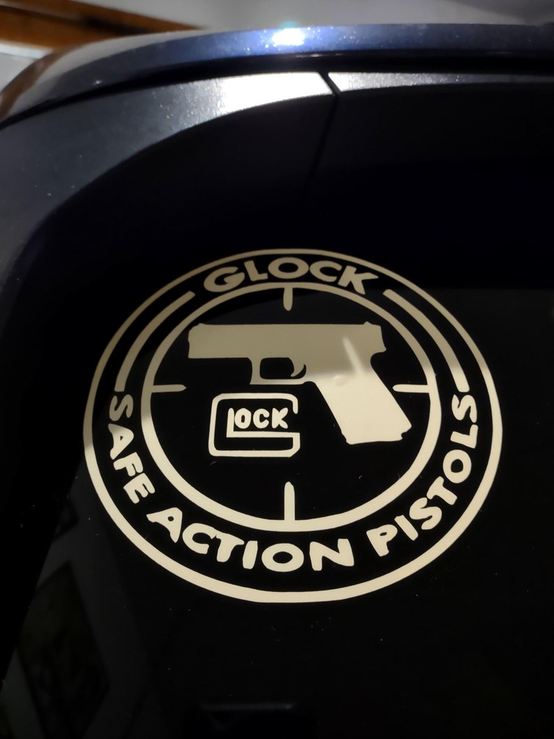 K&ouml;nnte beinhalten: Wei&szlig;er runder Aufkleber mit dem Wort "GLOCK" oben, einer Pistollensilhouette in der Mitte und den Worten "SAFE ACTION PISTOLS" unten. Der Aufkleber befindet sich auf einer dunkelblauen Oberfl&auml;che.