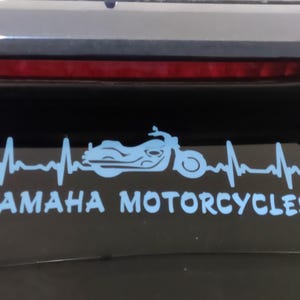 K&ouml;nnte beinhalten: Hellblauer Yamaha Motorcycles Aufkleber mit einer Motorradsilhouette in einer Herzschlaglinie. Der Text "YAMAHA MOTORCYCLES" befindet sich darunter. Der Aufkleber befindet sich auf einer dunklen Oberfl&auml;che.
