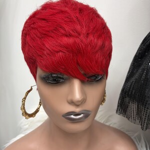 May include: A mannequin head wearing a short, vibrant red pixie cut wig with textured layers and side-swept bangs. The mannequin features metallic silver lipstick and large, gold-toned bamboo-style hoop earrings on both sides of the face.