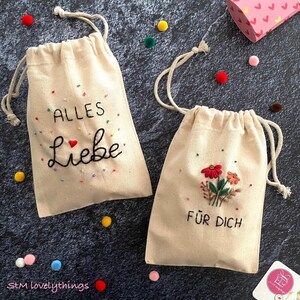 Könnte beinhalten: Zwei beigefarbene Kordelzugbeutel mit gestickten Designs. Auf einem Beutel stehen die Worte "ALLES Liebe" und auf dem anderen "FÜR DICH". Beide Beutel haben farbenfrohe Stickereiakzente und sind von bunten Pompons umgeben.