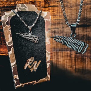May include: Two silver necklaces with feather pendants. The necklaces are displayed on a camouflage background. The pendant on the left is a detailed feather design. The pendant on the right is a more stylized feather design. The necklaces are both on a silver chain.