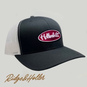 May include: Black trucker hat with a white mesh back and a curved brim. The front features an oval patch with the word "Hollenbilt" embroidered in white on a burgundy background. The hat has a snapback closure.