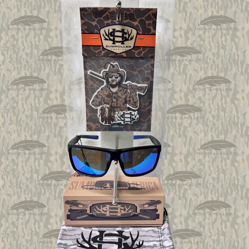 May include: Black sunglasses with blue mirrored lenses are displayed with packaging featuring a cowboy graphic and the text "SUMHOLLER AMERICA". The sunglasses have a rectangular frame and blue accents.